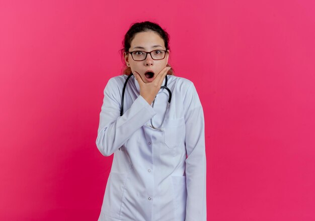 Doctora joven sorprendida vistiendo bata médica y estetoscopio y gafas sosteniendo la barbilla aislada en la pared rosa con espacio de copia