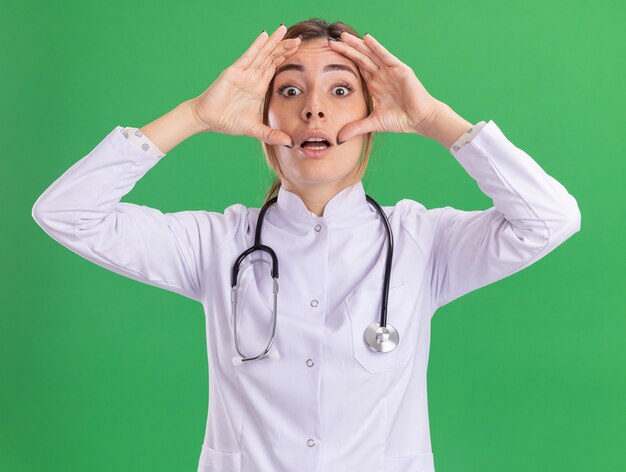 Doctora joven sorprendida vistiendo bata médica con estetoscopio poniendo las manos alrededor de los ojos aislados en la pared verde