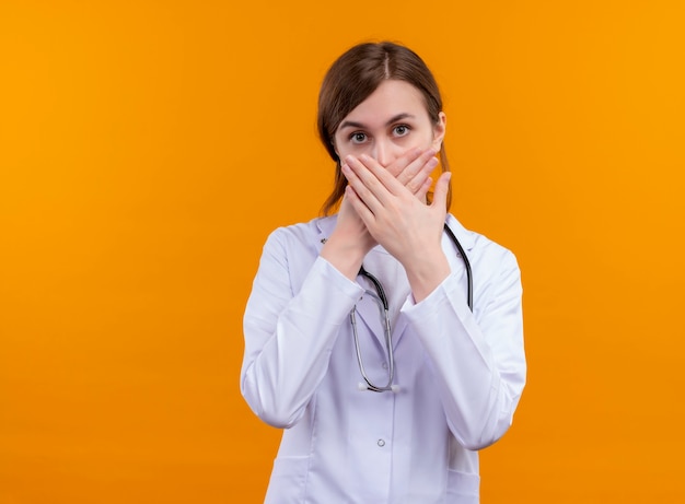 Doctora joven sorprendida vistiendo bata médica y estetoscopio poniendo las manos en la boca en la pared naranja aislada con espacio de copia