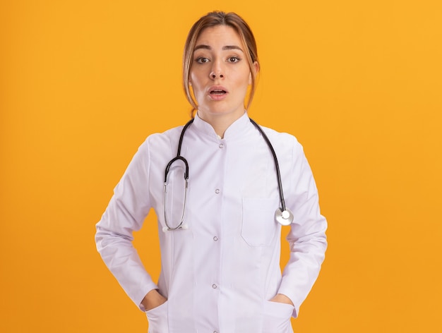 Doctora joven sorprendida vistiendo bata médica con estetoscopio poniendo las manos en el bolsillo aislado en la pared amarilla