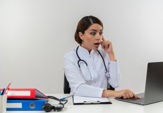 Doctora joven sorprendida vistiendo bata médica con estetoscopio sentado en el escritorio de trabajo en la computadora con herramientas médicas portátil usado poniendo el dedo en la mejilla con espacio de copia