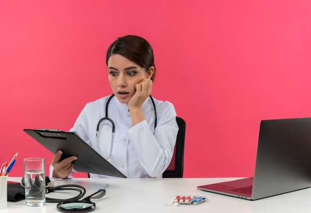 Doctora joven sorprendida vistiendo bata médica con estetoscopio sentado en el escritorio de trabajo en la computadora con herramientas médicas sosteniendo y mirando el portapapeles con espacio de copia