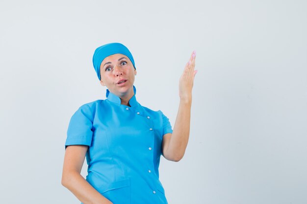 Doctora levantando el brazo y la palma en uniforme azul y mirando confundido. vista frontal.