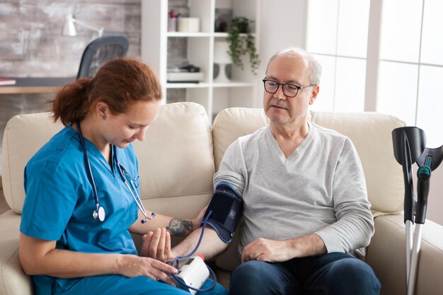 Doctora leyendo la presión arterial de un anciano en un hogar de ancianos usando un dispositivo digital. Enfermera y paciente sentados en el sofá.
