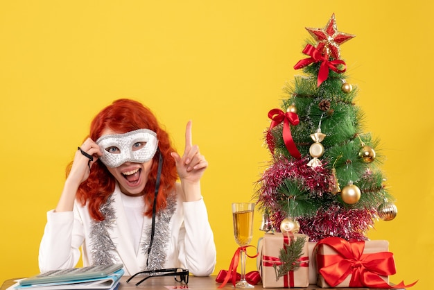 Doctora con máscara alrededor del árbol de Navidad y regalos en amarillo