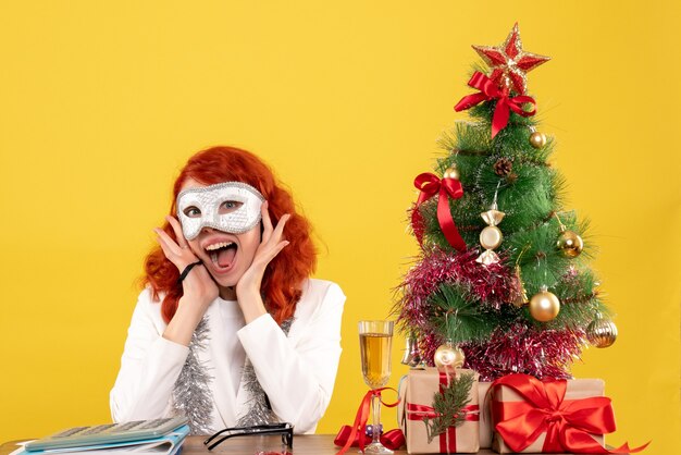 Doctora con máscara alrededor del árbol de Navidad y regalos en amarillo