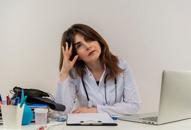 Doctora de mediana edad aburrida vistiendo bata médica y estetoscopio sentado en el escritorio con portapapeles de herramientas médicas y portátil tocando la cabeza mirando aislado
