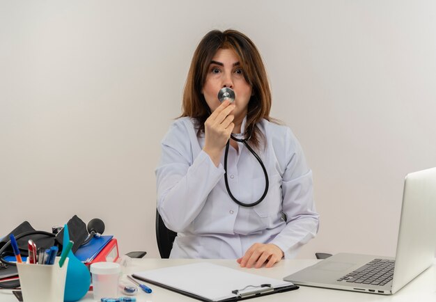 Doctora de mediana edad asustada vistiendo bata médica con estetoscopio sentado en el escritorio de trabajo en la computadora portátil con herramientas médicas poniendo estetoscopio en la boca en la pared blanca