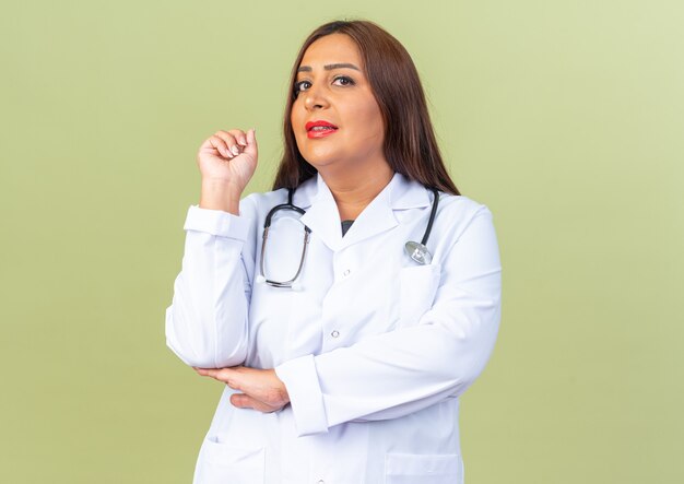 Doctora de mediana edad en bata blanca con estetoscopio con expresión escéptica apuntando hacia atrás de pie sobre la pared verde