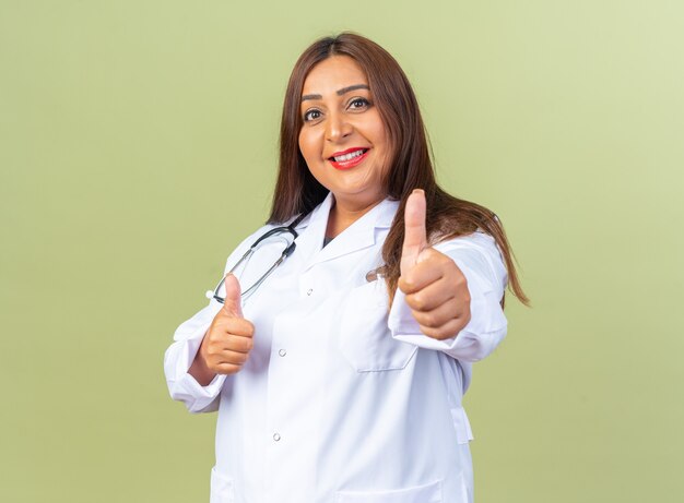 Doctora de mediana edad en bata blanca con estetoscopio feliz y positivo sonriendo alegremente mostrando los pulgares para arriba de pie sobre la pared verde