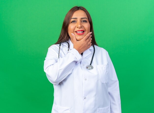 Doctora de mediana edad en bata blanca con estetoscopio feliz y positivo sonriendo alegremente de pie sobre la pared verde