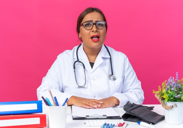 Doctora de mediana edad en bata blanca con estetoscopio con gafas confundido y muy ansioso sentado en la mesa sobre la pared rosa