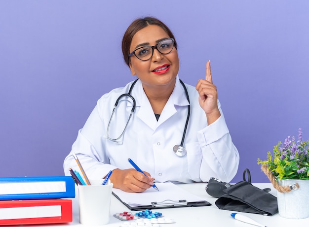 Doctora de mediana edad en bata blanca con estetoscopio con gafas mirando al frente sonriendo confiado mostrando el dedo índice con nueva idea sentado en la mesa sobre la pared azul