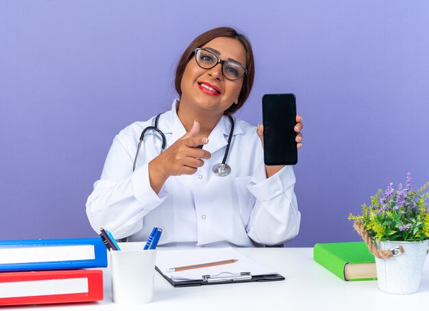 Doctora de mediana edad en bata blanca con estetoscopio con gafas mostrando smartphone apuntando con el dedo índice al frente sonriendo sentado en la mesa sobre la pared azul