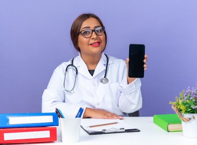 Doctora de mediana edad en bata blanca con estetoscopio con gafas mostrando smartphone sonriendo confiado sentado en la mesa sobre la pared azul
