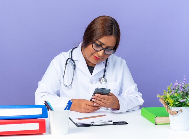 Doctora de mediana edad en bata blanca con estetoscopio con gafas sosteniendo smartphone mirándolo con una sonrisa en la cara sentado en la mesa sobre la pared azul