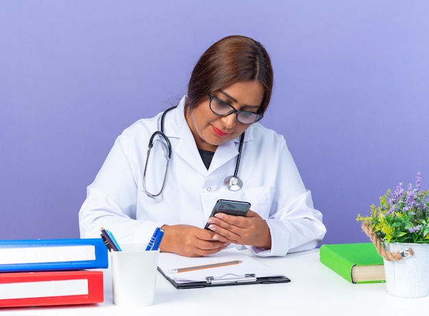 Doctora de mediana edad en bata blanca con estetoscopio con gafas sosteniendo smartphone mirándolo con una sonrisa en la cara sentado en la mesa sobre la pared azul