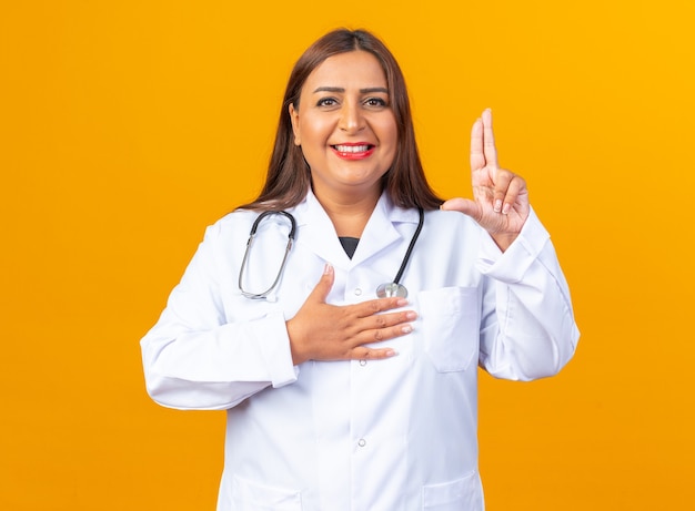 Doctora de mediana edad en bata blanca con estetoscopio haciendo gesto de juramento sonriendo confiado de pie sobre la pared naranja