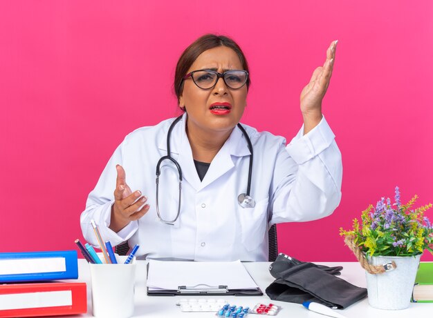 Doctora de mediana edad en bata blanca con estetoscopio mirando al frente confundido levantando los brazos con disgusto e indignación sentado en la mesa sobre la pared rosa