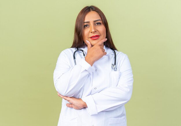 Doctora de mediana edad en bata blanca con estetoscopio mirando al frente con la mano en la barbilla pensando de pie sobre la pared verde