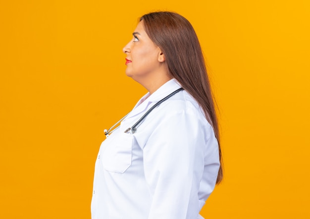 Doctora de mediana edad en bata blanca con estetoscopio mirando confiado de pie lateralmente en naranja