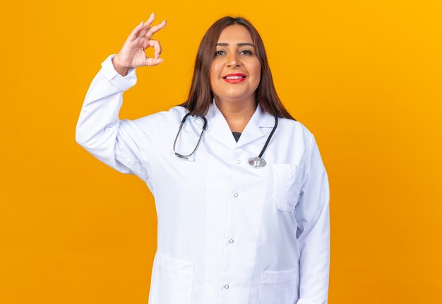 Doctora de mediana edad en bata blanca con estetoscopio mirando feliz y positivo sonriendo confiado mostrando signo ok