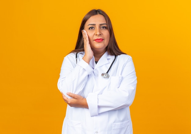 Doctora de mediana edad en bata blanca con estetoscopio mirando feliz y positivo sonriendo con la mano en el rostro