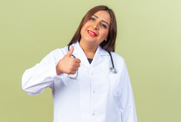 Doctora de mediana edad en bata blanca con estetoscopio mirando sonriendo confiado mostrando los pulgares para arriba