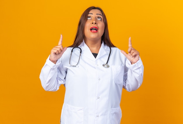 Doctora de mediana edad en bata blanca con estetoscopio mirando sorprendido apuntando con los dedos índices de pie sobre la pared naranja