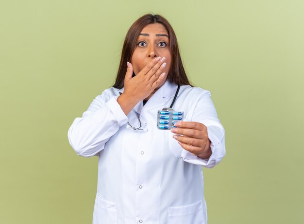 Doctora de mediana edad en bata blanca con estetoscopio mostrando blister con pastillas que está sorprendido cubriendo la boca con la mano de pie sobre la pared verde
