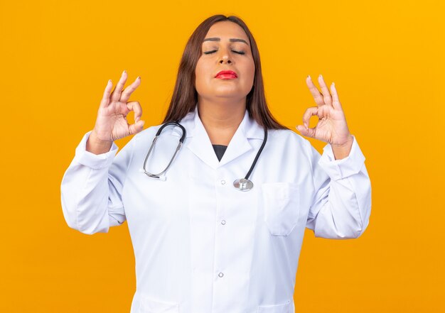 Doctora de mediana edad en bata blanca con estetoscopio relajante haciendo gesto de meditación con los dedos con los ojos cerrados