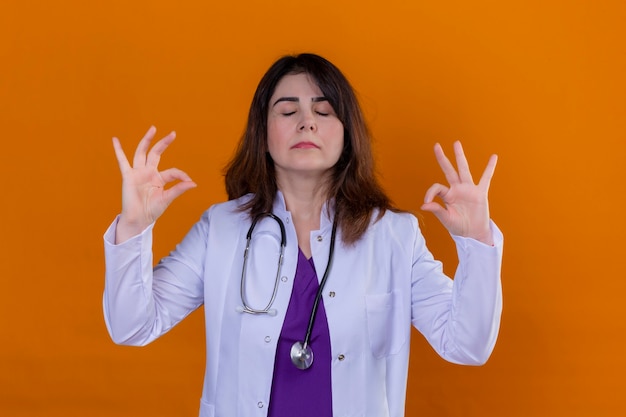 Foto gratuita doctora de mediana edad con bata blanca y con estetoscopio relajarse y sonreír con los ojos cerrados haciendo gesto de meditación con los dedos sobre fondo naranja
