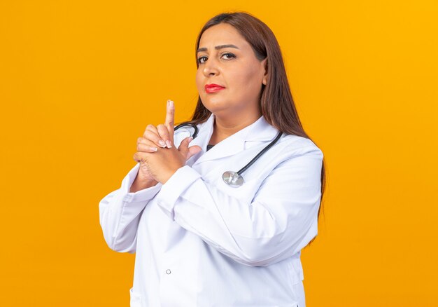 Doctora de mediana edad en bata blanca con estetoscopio con rostro serio haciendo gesto de pistola con los dedos
