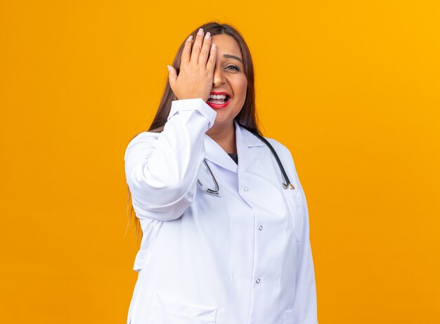 Doctora de mediana edad en bata blanca con estetoscopio sonriendo alegremente cubriendo los ojos con la mano de pie sobre la pared naranja