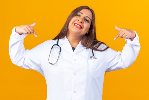 Doctora de mediana edad en bata blanca con estetoscopio sonriendo confiado, feliz y positivo apuntando a sí mismo de pie sobre la pared naranja