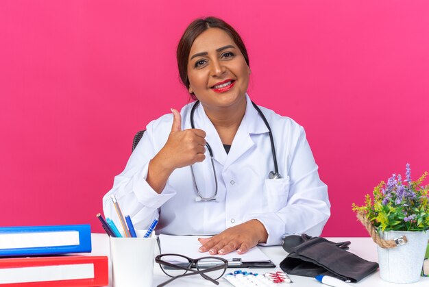 Doctora de mediana edad en bata blanca con estetoscopio sonriendo confiado mostrando los pulgares para arriba sentado en la mesa con carpetas de oficina en rosa