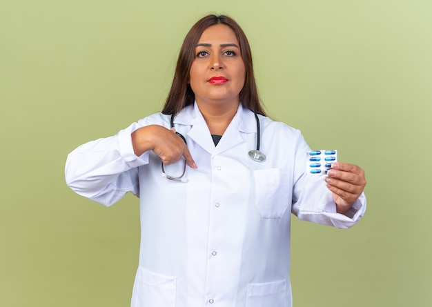 Doctora de mediana edad en bata blanca con estetoscopio sosteniendo blister con pastillas mirando con expresión de confianza apuntando a sí misma