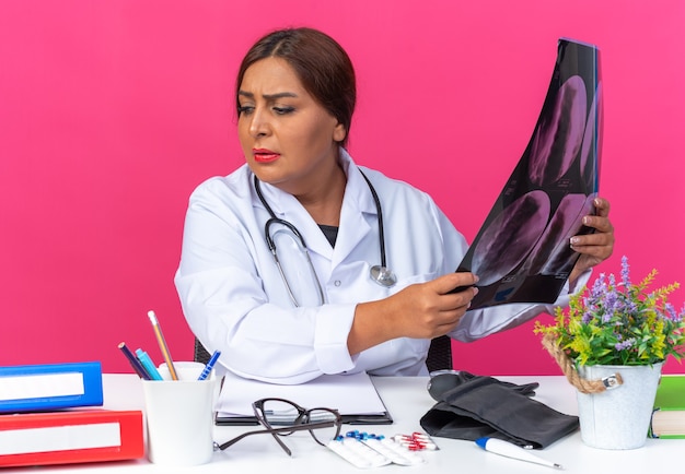 Doctora de mediana edad en bata blanca con estetoscopio sosteniendo rayos x mirando a un lado confundido y muy ansioso sentado en la mesa con carpetas de oficina en rosa