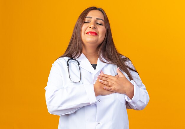 Doctora de mediana edad en bata blanca con estetoscopio tomados de la mano en el pecho sintiendo emociones positivas de pie sobre la pared naranja