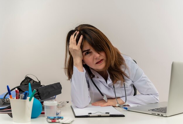 Doctora de mediana edad cansada con bata médica y estetoscopio sentado en el escritorio con portapapeles de herramientas médicas y computadora portátil poniendo la mano en la cabeza mirando aislado