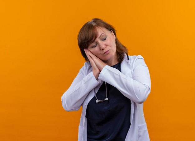 Doctora de mediana edad cansada vistiendo bata médica y estetoscopio poniendo la cabeza en las manos en la pared naranja aislada con espacio de copia