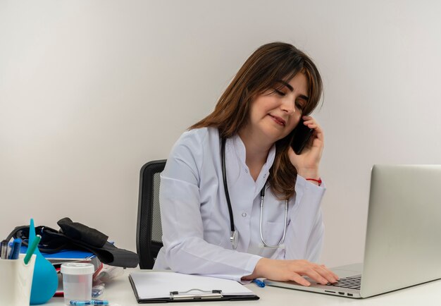 Doctora de mediana edad complacida con bata médica y estetoscopio sentado en el escritorio con portapapeles de herramientas médicas y computadora portátil hablando por teléfono mirando hacia abajo usando una computadora portátil aislada