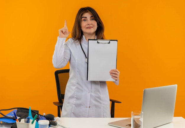 Doctora de mediana edad complacida con bata médica con estetoscopio sentado en el escritorio trabajando en una computadora portátil con herramientas médicas sosteniendo el portapapeles y apunta hacia arriba en la pared naranja con espacio de copia