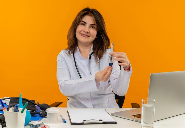 Doctora de mediana edad complacida con bata médica con estetoscopio sentado en el escritorio trabajando en una computadora portátil con herramientas médicas sosteniendo un termómetro en una pared naranja aislada con espacio de copia