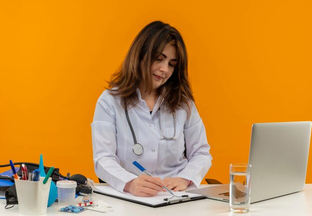 Doctora de mediana edad complacida con bata médica con estetoscopio sentado en el escritorio, trabajo en una computadora portátil con herramientas médicas, escriba en el portapapeles en una pared naranja aislada con espacio de copia