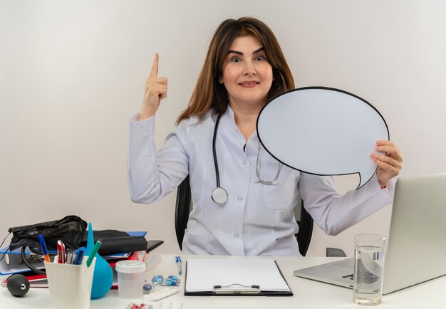 Foto gratuita doctora de mediana edad complacida con bata médica con estetoscopio sentado en el escritorio, trabajo en una computadora portátil con herramientas médicas que sostienen la burbuja de chat y apunta hacia arriba en la pared blanca aislada