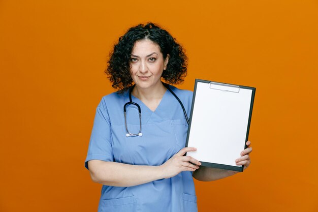 Doctora de mediana edad complacida con uniforme y estetoscopio alrededor del cuello mirando a la cámara que muestra el portapapeles aislado en el fondo naranja