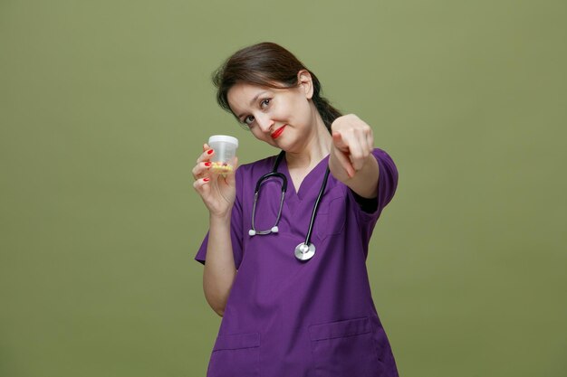 Doctora de mediana edad complacida con uniforme y estetoscopio alrededor del cuello que muestra un recipiente de medición con pastillas mirando a la cámara apuntando a la cámara aislada en un fondo verde oliva