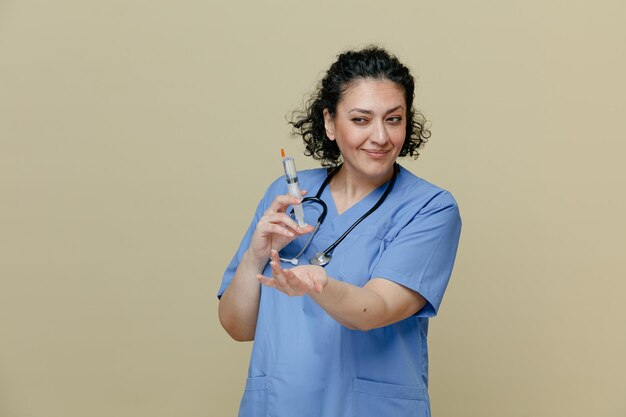 Una doctora de mediana edad complacida con uniforme y estetoscopio alrededor del cuello sosteniendo una jeringa con una aguja mirando al costado mostrando un gesto de venir aquí aislado en un fondo verde oliva