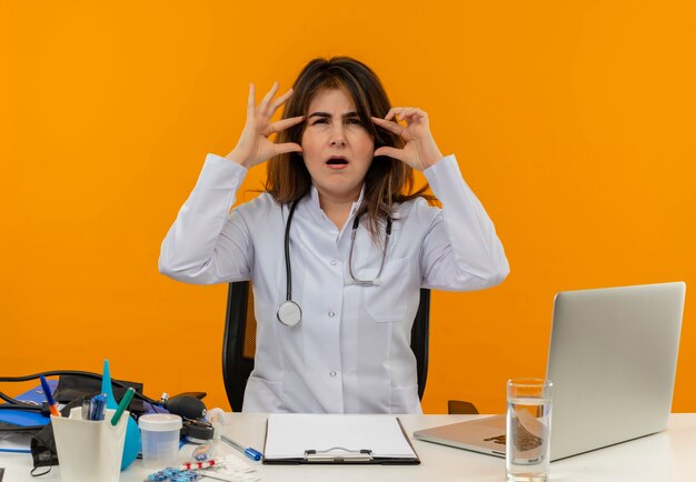 Doctora de mediana edad confundida con bata médica con estetoscopio sentado en el escritorio, trabajo en una computadora portátil con herramientas médicas, poniendo los dedos en la frente en la pared naranja aislada con espacio de copia